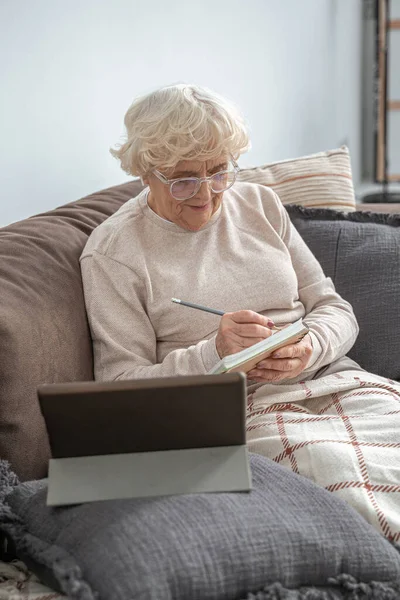 Зріла жінка в окулярах сидить на дивані вдома і використовує планшет — стокове фото