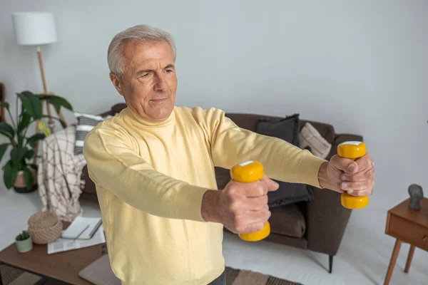 Senior trainiert zu Hause mit Kurzhanteln, während er wegschaut und sich glücklich fühlt — Stockfoto