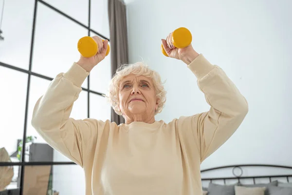 Ältere Frau im Rentenalter trainiert zu Hause mit orangefarbenen Hanteln und lächelt — Stockfoto
