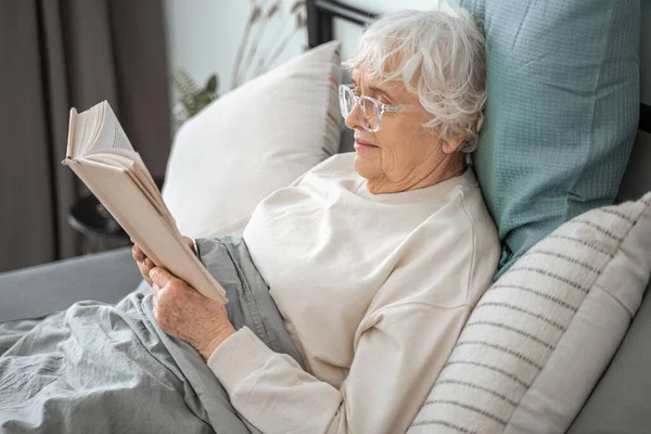 긴장 한 성숙 한 여자가 침대에 앉아 가장 좋아 하는 책을 읽고 있다 — 스톡 사진