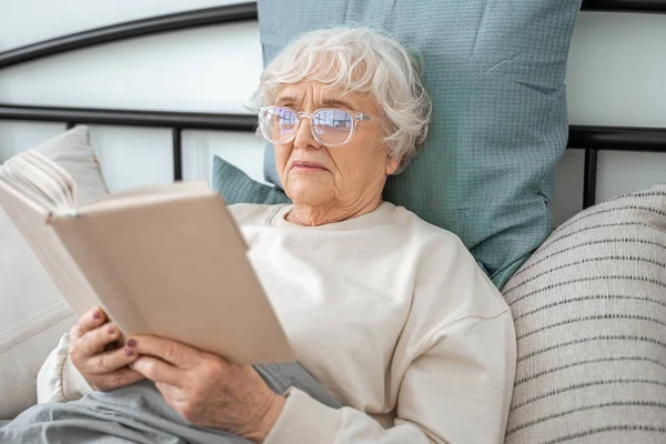 Старша жінка тримає книгу, читаючи свій улюблений роман вдома — стокове фото