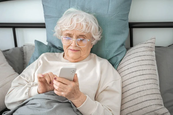 Усміхнена зріла жінка дивиться на мобільний телефон, лежачи на затишному ліжку — стокове фото