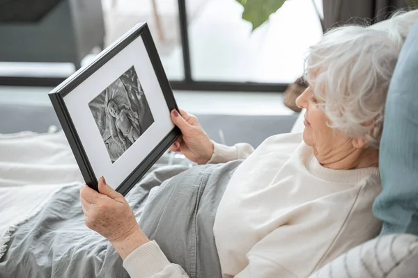 Mujer mayor mirando la foto en el marco mientras se relaja en la cama —  Fotos de Stock