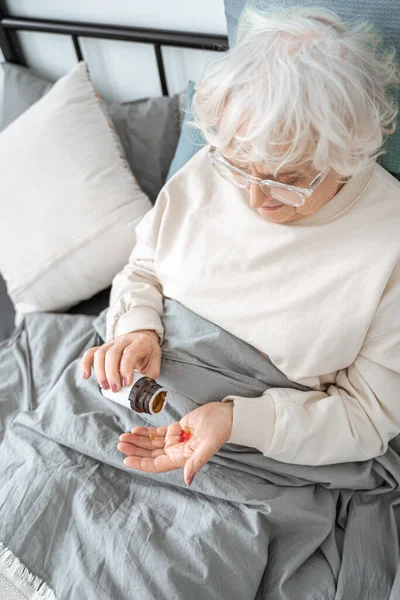 Yatakta otururken elinde haplı kavanoz tutan emekli kadın. — Stok fotoğraf