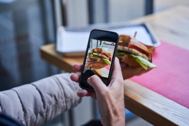 Elderly man sitting at the table at the cafe and shooting tasty sandwich