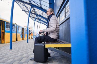 Mature man with black suitcase sitting on bench at bus stop while waiting for the bus