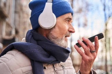 Senior guy wearing headphones recording voice message at his smartphone