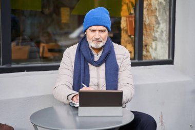 Mature man writing some notes and looking at the tablet computer while working