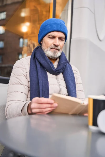 Homme âgé en vêtements chauds assis avec un livre à la terrasse du café — Photo