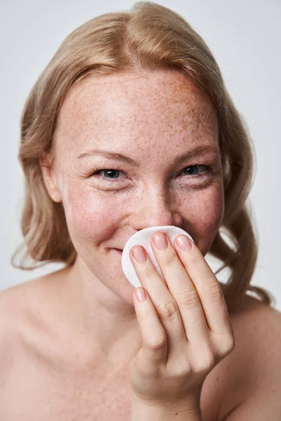 Mujer pecosa sonriente escondiendo su cara detrás de la almohadilla de algodón mientras se quita el maquillaje —  Fotos de Stock