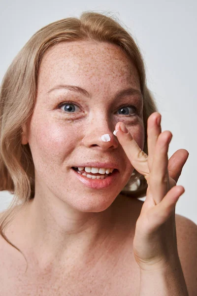 Роздягнена жінка кладе зволожуючий крем на ніс і посміхається — стокове фото