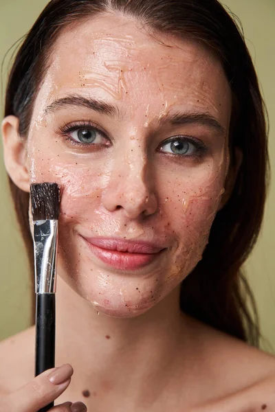 Mujer aplicando mascarilla de arcilla para el rejuvenecimiento de la piel mientras sostiene cepillo cosmético —  Fotos de Stock