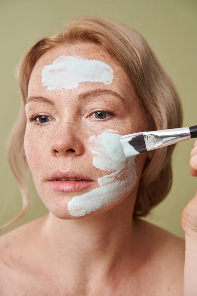 Mujer aplicando máscara natural azul con cepillo cosmético y mirando hacia otro lado —  Fotos de Stock