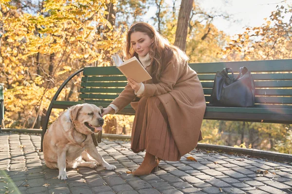 Femme adulte souriante caressant son chien et lisant un livre sur le banc — Photo