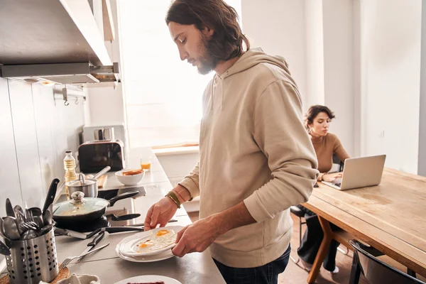 Man förbereder frukost i köket medan hans fru använder laptop — Stockfoto