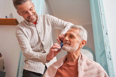 Oğlu sakalını nazikçe keserken elinde tarak ve bukle tutarken gülen mutlu beyaz adam.