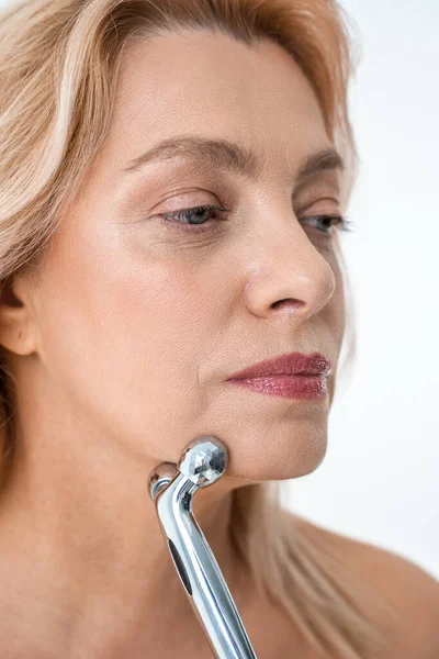 Mujer rubia mayor mirando hacia otro lado con herramienta de masaje facial sobre fondo blanco — Foto de Stock