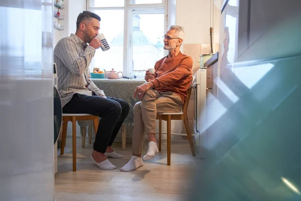 Senior vader draagt bril praten en het drinken van koffie met zijn volwassen zoon in de keuken — Stockfoto