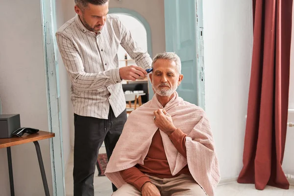 Concentrated adult man making haircut for his father at home with haircut machine