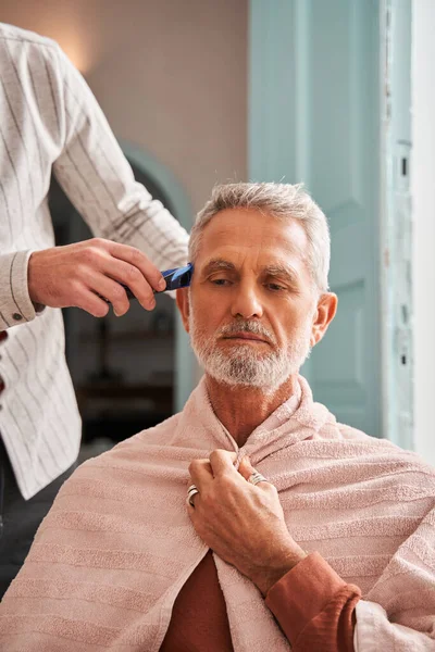 정신 집중이 된 아들 이 빗자루와 재단기를 사용하면서 은퇴 한 아버지에게 머리를 깎고 있는 모습 — 스톡 사진