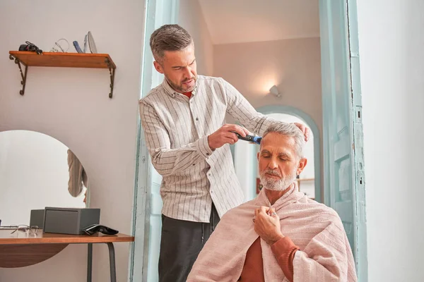 Volwassen man met baard knipt zijn vader in de woonkamer voor de spiegel — Stockfoto