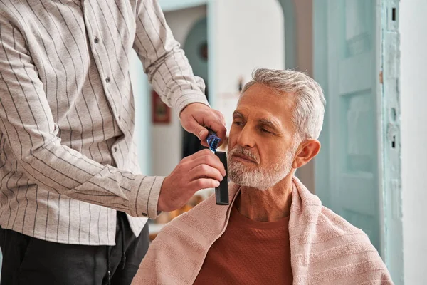 Grijs harige man genieten terwijl zijn zoon snijden zijn baard met kam en trimer — Stockfoto