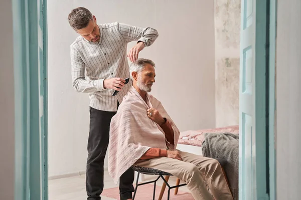 Oudere knappe man krijgt zijn haar en baard geknipt thuis terwijl zijn zorgvuldige zoon te helpen — Stockfoto