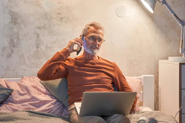 Glimlachende oudere man in huishoudelijke kleding met laptop en het maken van een telefoontje met de smartphone — Stockfoto