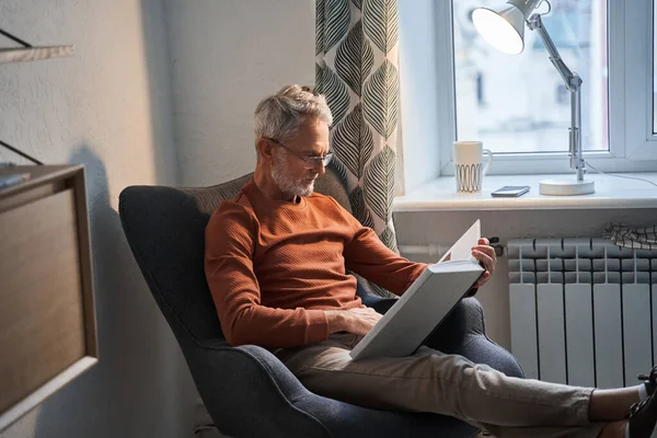 Senior man draagt een bril ontspannen in de fauteuil thuis tijdens het lezen van zijn favoriete boek — Stockfoto