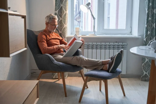 Biały senior relaksujący się na fotelu w domu czytając swoją ulubioną książkę — Zdjęcie stockowe