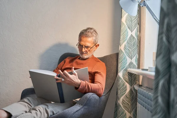 Ernstige ongeschoren gepensioneerde man lezen interessant boek terwijl situeren op gezellige fauteuil in appartement — Stockfoto