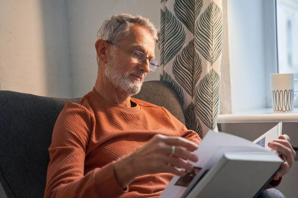 Glad farfar läser bok medan du tittar igenom glasögonen. Han sitter hemma i fåtöljen. — Stockfoto