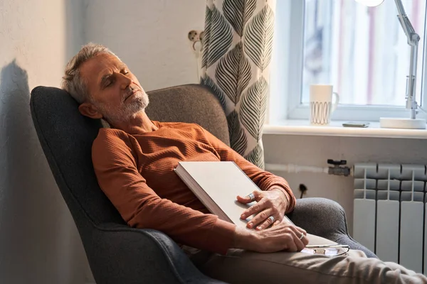 Втомився зрілий чоловік спить біля крісла з книгою на грудях після читання — стокове фото