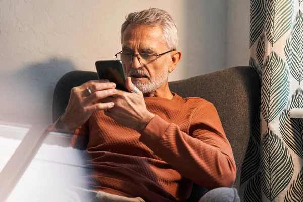 Glimlachende bejaarde man thuis met moderne smartphone — Stockfoto