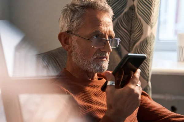 Mature old man using smartphone while relaxing on sofa and chatting with somebody — Stock Photo, Image