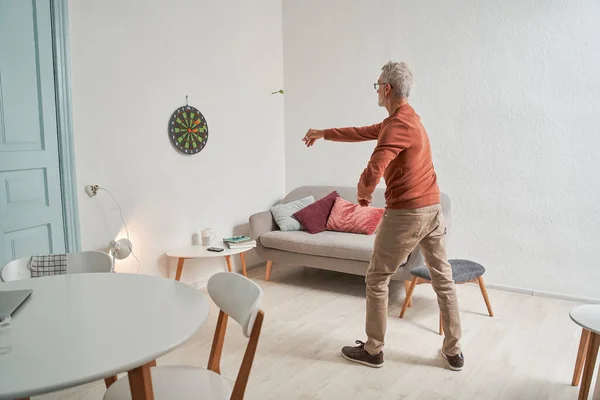Senior Kaukasische man gooien darts in gezellige kamer interieur, terwijl spelen alleen thuis — Stockfoto