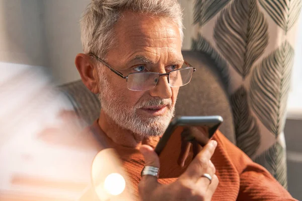 Gammal man spela in röstmeddelande via smartphone när du sitter vid fåtöljen nära fönstret — Stockfoto