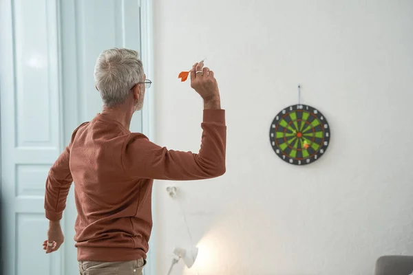 Gris pelo maduro hombre jugando dardos en casa en su acogedora habitación —  Fotos de Stock