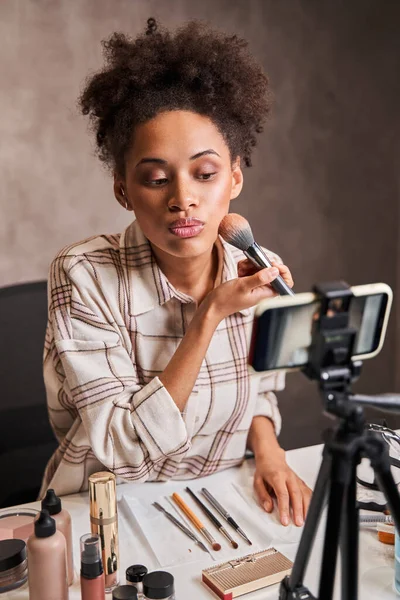Attentive curly multiracial woman applying makeup on her face while streaming — Stock Photo, Image