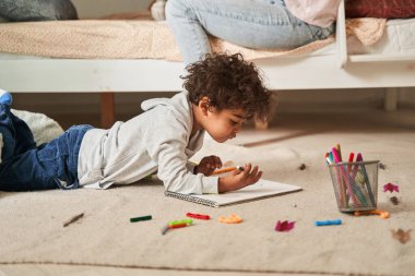 Küçük çok kültürlü çocuk yerde yatıyor ve kağıda kurşun kalemlerle bir şeyler çiziyor.