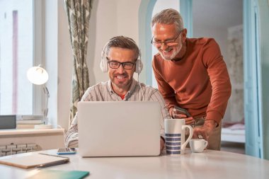 Neşeli yaşlı baba, yetişkin oğlu çalışırken bilgisayarın ekranına bakıp gülümsüyor.
