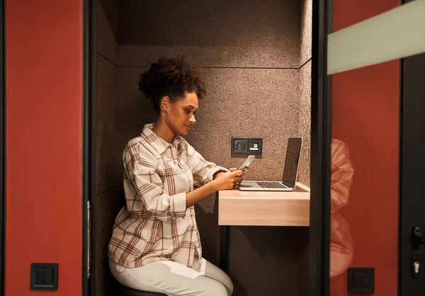 Frau sitzt während der Arbeit mit Laptop und Smartphone in kleinem Raum am Tisch — Stockfoto