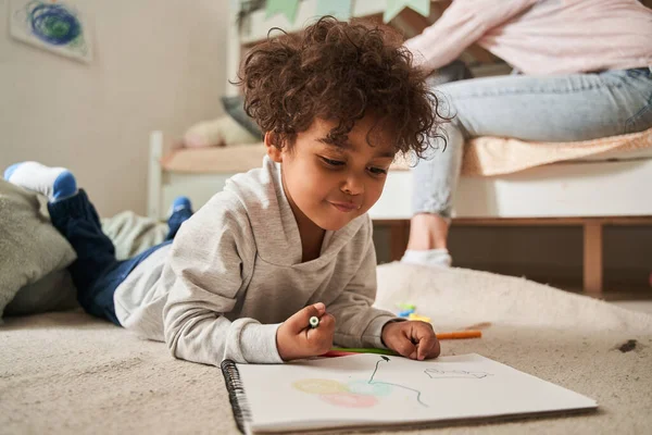 Pequeno menino multirracial deitado no chão e desenhando algo com lápis no papel — Fotografia de Stock