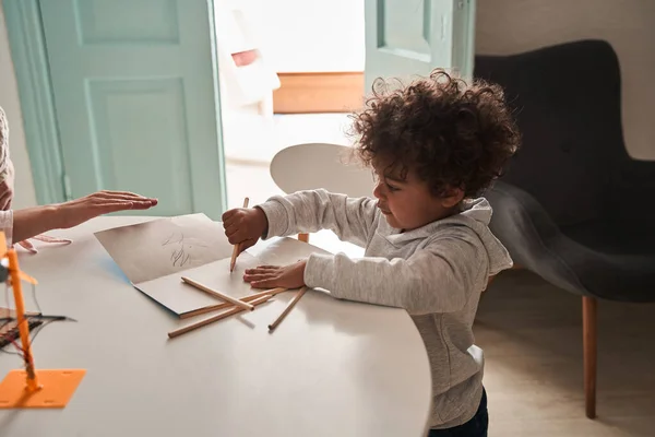 Morena encaracolado menino de pé à mesa e desenho enquanto passa o tempo na cozinha — Fotografia de Stock