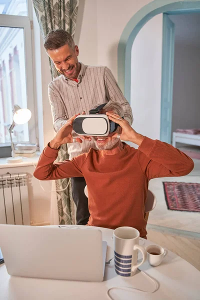 Pria Kaukasia dewasa yang tampan menyesuaikan headset VR-nya dengan senyuman manis pada seniornya — Stok Foto