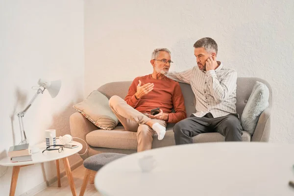 Pria pensiunan dengan anaknya yang sudah dewasa bersantai di sofa di apartemen mereka yang nyaman — Stok Foto