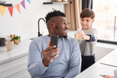 Baba ve oğul akıllı telefon kamerasına ateş ederken birbirlerine bakıyorlar.