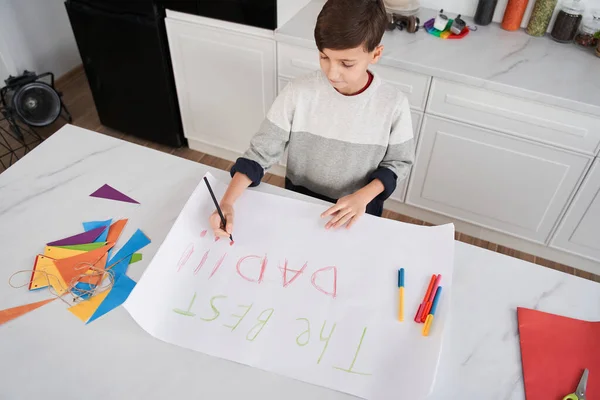 Beyaz çocuk masada oturuyor ve evde babası için pankart çiziyor. — Stok fotoğraf