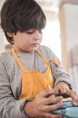 Önlüklü beyaz çocuk masada duruyor ve elinde ciddi yüzlü boyalarla kavanozu tutuyor.