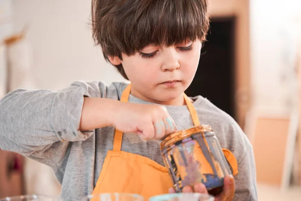 Boy wearing apron standing at the table and holding jar with paints while making drawings — Stock Photo, Image